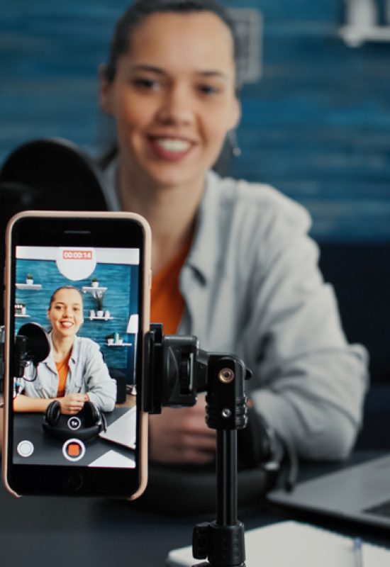 Attractive social media influencer sitting at home studio desk while starting streaming from living room with smartphone. Young content creator recording daily vlog with touchscreen modern device.